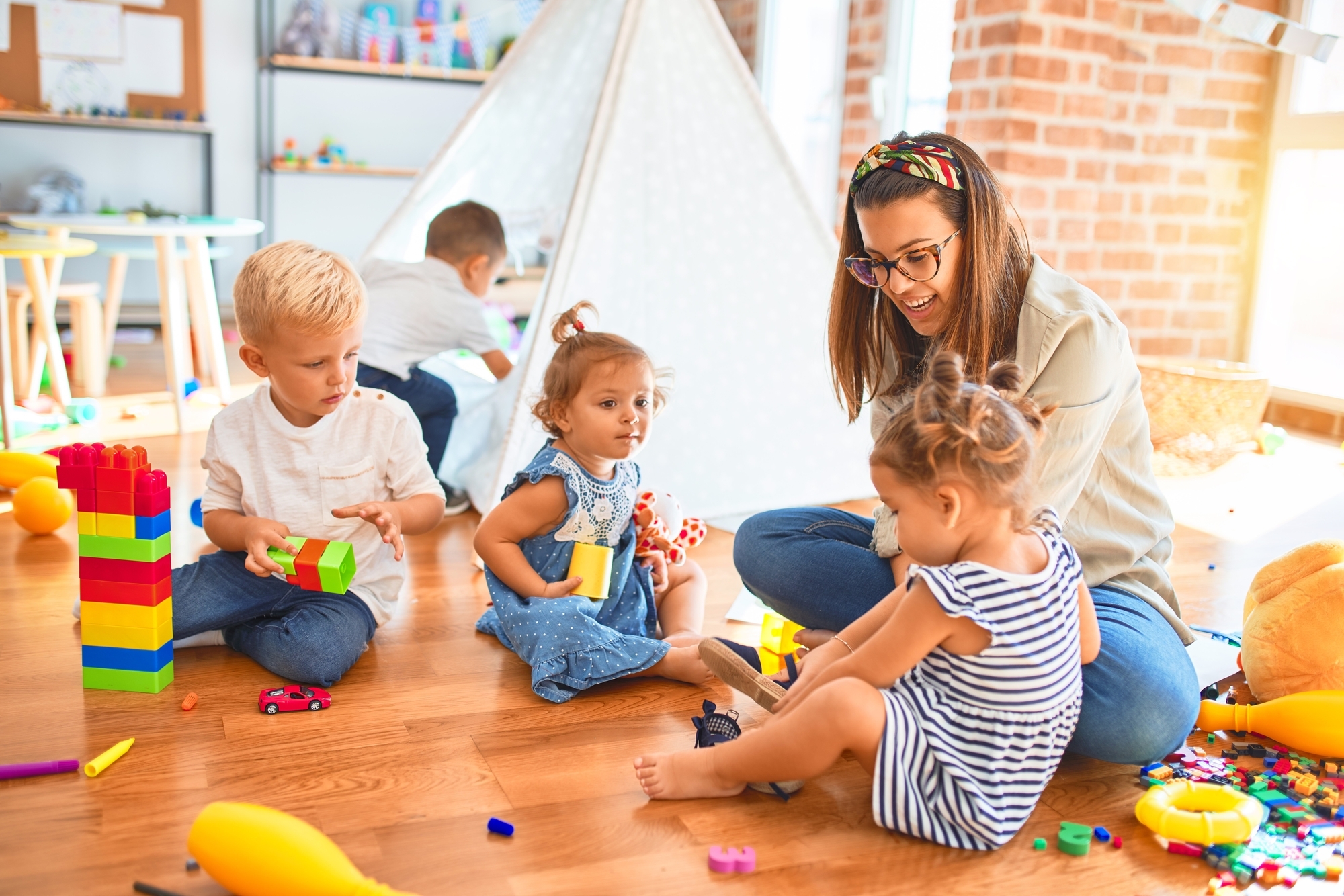 Beautiful teacher and group of toddlers playing around lots of toys at kindergarten