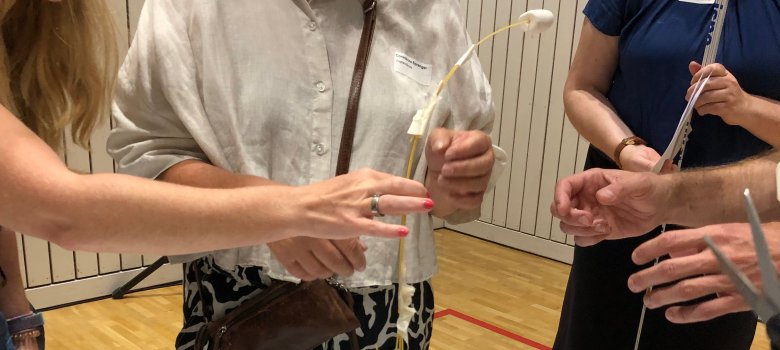Hände über einm Tisch, die versuchen einen Marshmallow auf einer Nudel zu platzieren