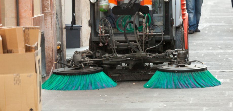 Eine Straße an der ein Gebäude angrenzt, wird von einer Straßenkehrmaschine mit zwei Bürstenaufsätzen gereinigt.