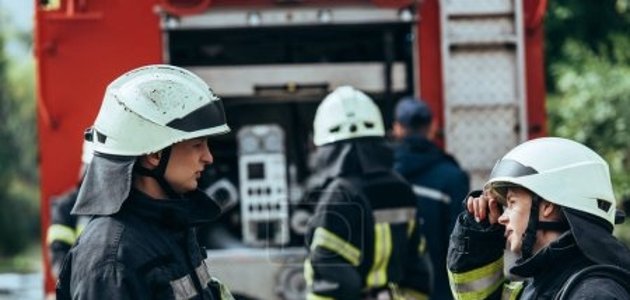 Zusehen ist hintergründig ein Einsatzfahrzeug der Feuerwehr. Davor stehen zwei Feuerwehrleute in Einsatzkleidung.