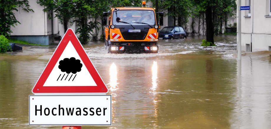 Auf einer überschwemmten Straße versucht ein orangenes Einsatzfahrzeug diese zu durchqueren. Im vorderen unteren Bereich des Bildes ist ein dreieckiges Verkehrsschild mit einer Gewitterwolke und der Zusatzbezeichnung "Hochwasser" zusehen.