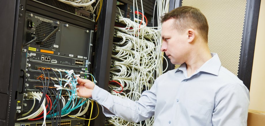 Männliche Person mit blauem Hemd steht vor einem Hardware-Server-Turm und kontrolliert die einzelnen Anschlüsse.